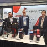 Three men in business attire stand behind a booth displaying coin and note recycling machines at a technology expo, part of the carwash trade show tour. A banner for Innovative Technology is visible behind them, along with other promotional materials.