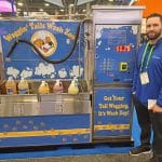 A man stands next to a self-service dog wash station labeled Waggin Tails Wash Zone, featuring soap bottles, a hose, and a $1.75 price sign at a carwash trade show tour.
