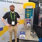 A man with a beard and glasses, wearing a black polo shirt and khaki pants, stands next to a yellow-topped vacuum machine at a carwash trade show booth. A sign invites people to visit booth 409 during the tour.