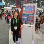A woman in a black shirt stands smiling next to a large touchscreen kiosk at a carwash trade show. The kiosk displays WashApp and various washing service options, while other attendees and booths are visible in the background.