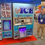 A man stands next to a large ice vending machine with Fresh Bagged ICE signs and prices for 10, 20, and 30 lbs. The booth is set up in an indoor exhibition space during a carwash trade show tour.