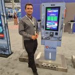 A man in business attire stands smiling beside a tall touchscreen kiosk labeled Colonel Clean at a carwash trade show booth. The kiosk displays a car wash menu and payment options, with informational displays highlighting the tour in the background.