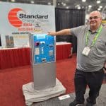 A man in a gray shirt and glasses stands smiling next to a change machine display at a carwash trade show. The booth behind him has a Standard Change-Makers banner and informational materials on a red and white table.