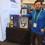 A man in a blue MONEX shirt stands next to a display of payment terminals and control panels at a carwash trade show booth. He is smiling and has one hand resting on the display table, ready to welcome visitors on the trade show tour.