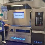 An elderly man smiles next to a large, high-tech dog washing machine labeled CYBERWASH 3000 at a carwash trade show. A screen on the machine displays the message, A CLEAN DOG IS A HAPPY DOG!.