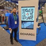 A man stands next to a QCharge electric vehicle charging station displaying a large QR code on its screen at a carwash trade show. The background features charging equipment and booths on a blue carpeted floor.