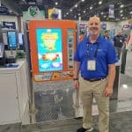 A smiling man in a blue shirt and khaki pants stands indoors next to a colorful, upright touchscreen kiosk with bright graphics, at a carwash trade show tour featuring other technology displays in the background.