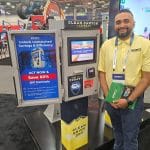 A smiling man in a yellow shirt stands next to a car wash payment kiosk with promotional signage offering 60% off controls at a carwash trade show tour booth.