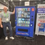 A man stands next to a blue Auto Vend Plus vending machine at a carwash trade show booth, with a promotional sign displaying price and special offers for attendees on the tour.