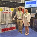 A woman and a man stand smiling in front of a large stainless steel machine labeled WATERMILL DOG WASH at a carwash trade show; banners and booths are visible in the background.