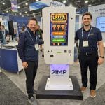 Two men stand beside a white kiosk with a Lucky 7s Car Wash slot machine display at a carwash trade show booth. Both are smiling, wearing badges, and the booth features banners and promotional materials in the background.
