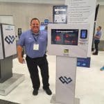 A smiling man in a blue shirt stands beside a touchscreen kiosk at a carwash trade show booth for WASHLINK. The booth displays various equipment and branding, while other attendees tour the exhibition in the background.