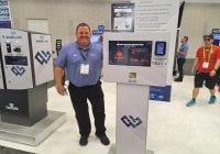A smiling man in a blue shirt stands beside a touchscreen kiosk at a carwash trade show booth for WASHLINK. The booth displays various equipment and branding, while other attendees tour the exhibition in the background.