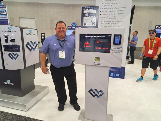 A smiling man in a blue shirt stands beside a touchscreen kiosk at a carwash trade show booth for WASHLINK. The booth displays various equipment and branding, while other attendees tour the exhibition in the background.