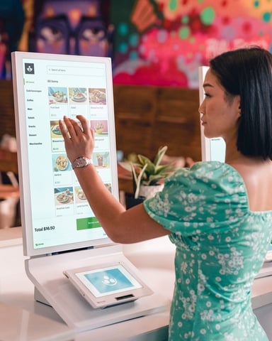 Clover kiosk Fiserv A woman in a green dress uses a Clover kiosk to order food in a colorful, modern restaurant. The touchscreen displays menu options and her total price.