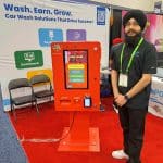 A man in a black turban and shirt stands next to a bright red digital kiosk at a carwash trade show tour booth promoting wash solutions. The screen displays options, with informational banners visible in the background.