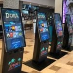 Four digital ticket kiosks for Palms Theatres & IMAX stand in a row inside a lobby, displaying movie options like Dora on their digital signage screens. A bar and dining area are visible in the background.